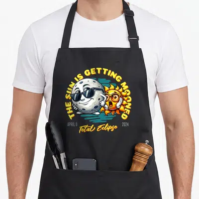 Getting april apron - Cartoon moon and sun with THE SUN IS GETTING MOONED Total Eclipse 2024 text — shown on a black cotton canvas apron against a white background
