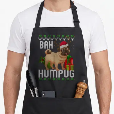 Bah xmas apron - Tan pug with antlers and red hat surrounded by gifts and BAH HUMPUG text — shown on a black cotton canvas apron against a white background