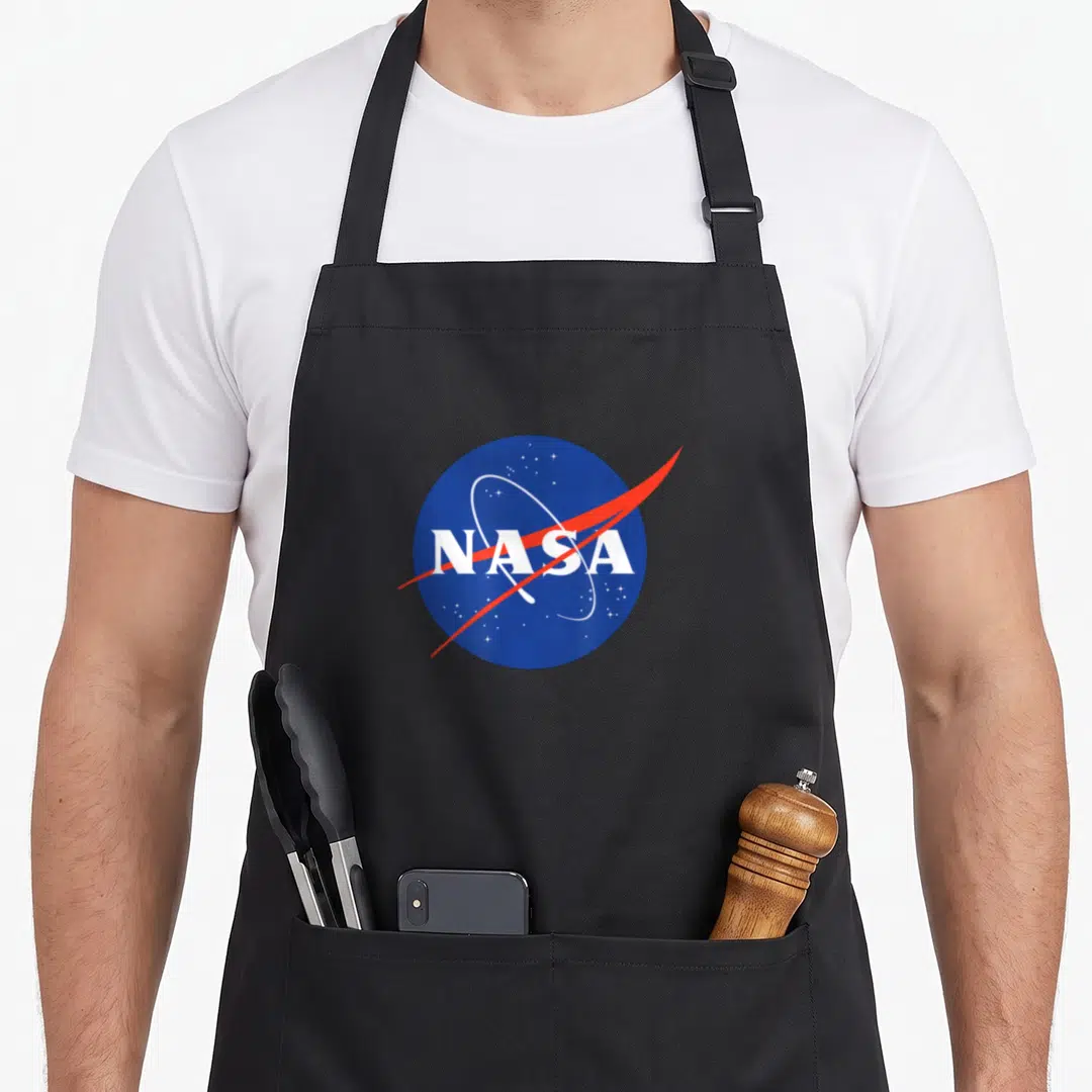 Cute meatball nasa apron - Blue circle with white NASA text, red swoosh, white orbit ring, and stars — shown on a black cotton canvas apron against a white background