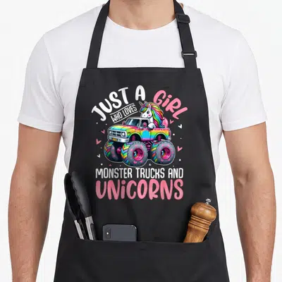 Unicorn toddler truck apron - Cartoon unicorn driving rainbow monster truck with text just a girl who loves monster — shown on a black cotton canvas apron against a white background