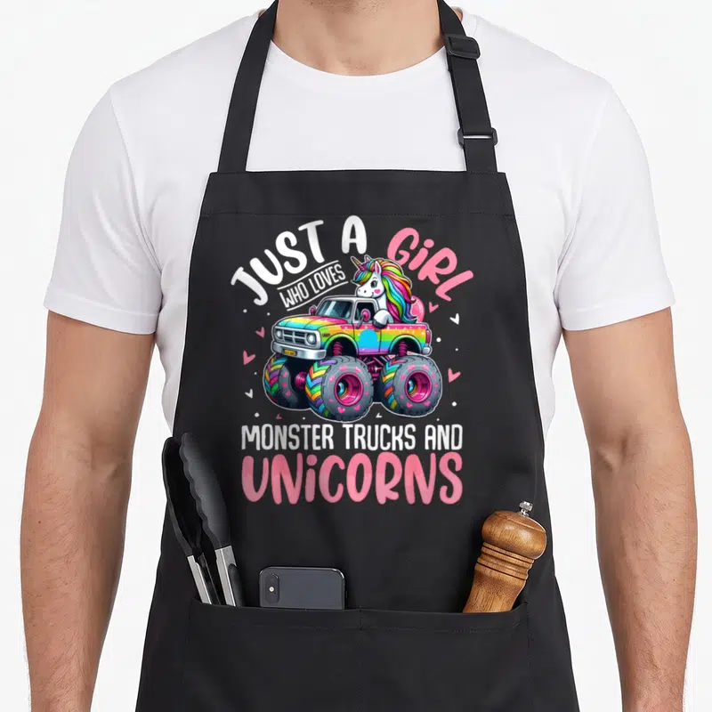 Unicorn toddler truck apron - Cartoon unicorn driving rainbow monster truck with text just a girl who loves monster — shown on a black cotton canvas apron against a white background