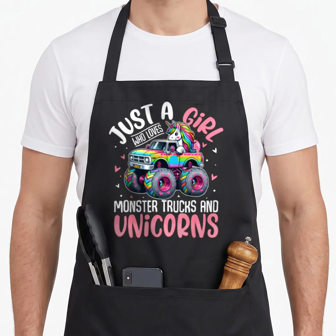 Unicorn toddler truck apron - Cartoon unicorn driving rainbow monster truck with text just a girl who loves monster — shown on a black cotton canvas apron against a white background