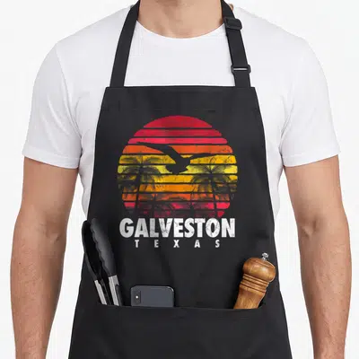Sunset texas apron - Striped red orange yellow sunset with bird palm trees and Galveston Texas text — shown on a black cotton canvas apron against a white background