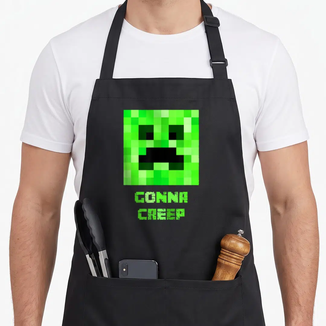 Creep face apron - Pixelated green creeper face above GONNA CREEP text in pixel font — shown on a black cotton canvas apron against a white background