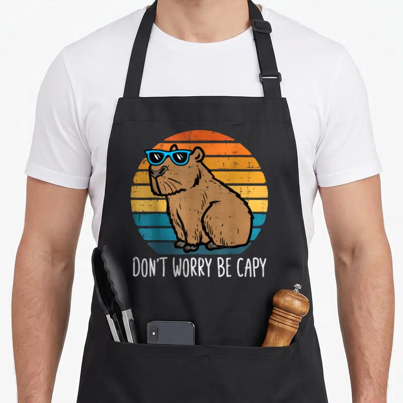 Worry cute capybara apron - Brown capybara in teal sunglasses before striped retro sun with white text DON'T WORRY BE — shown on a black cotton canvas apron against a white background
