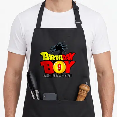 9th anime mom apron - Dragon head over BIRTHDAY BOY text with yellow 9 sphere and Japanese greeting — shown on a black cotton canvas apron against a white background