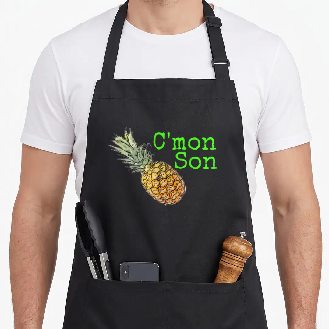 Cmon pineapple apron - Cartoon pineapple with stacked C'mon Son text in green font — shown on a black cotton canvas apron against a white background
