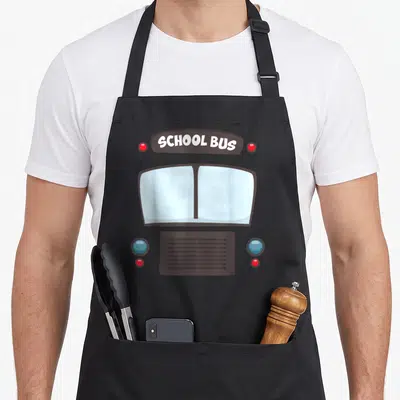 School bus omnibus apron - Cartoon bus front with brown sign reading SCHOOL BUS and blue and red lights — shown on a black cotton canvas apron against a white background
