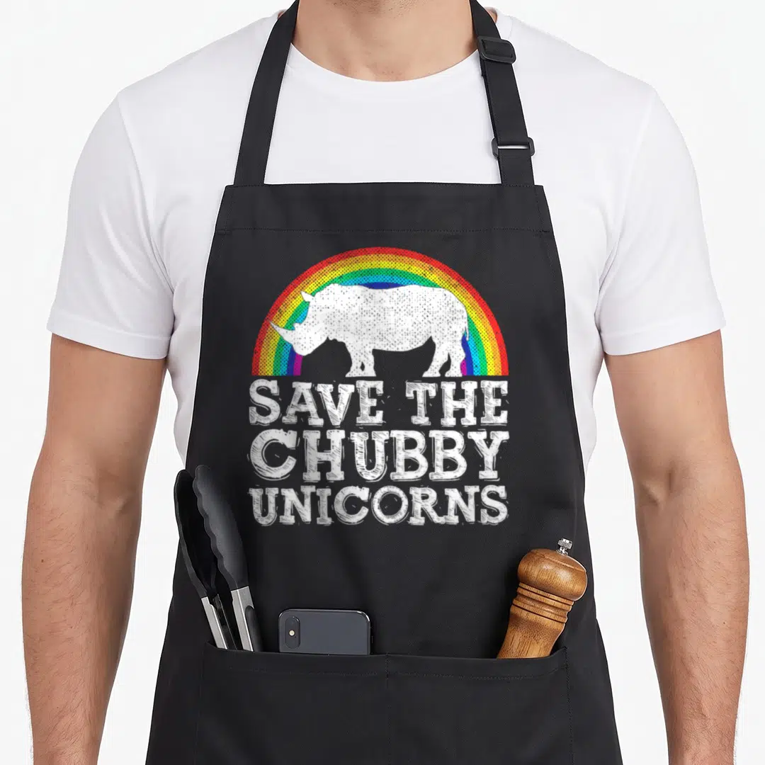 Save rainbow apron - White rhino silhouette under rainbow arch with distressed text SAVE THE CHUBBY UNICORNS — shown on a black cotton canvas apron against a white background