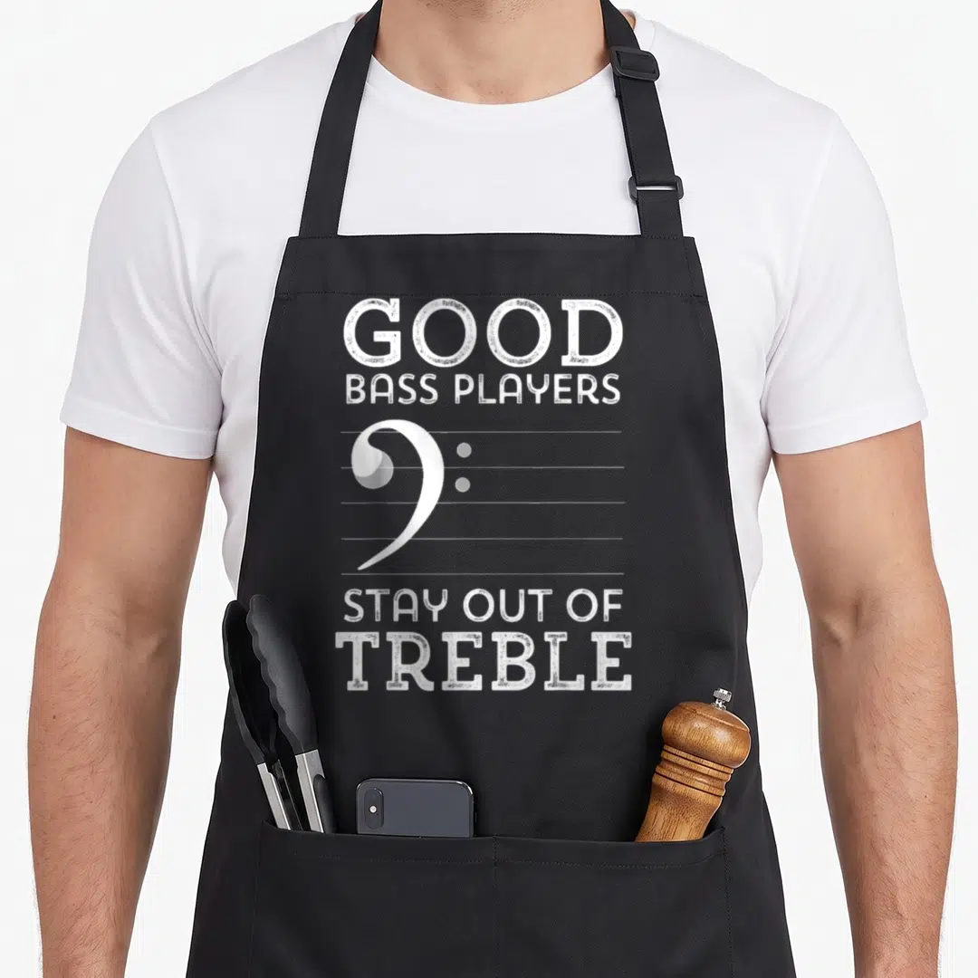 Stay bassoon apron - Good Bass Players Stay Out of Treble text surrounding a white bass clef on a musical staff — shown on a black cotton canvas apron against a white background