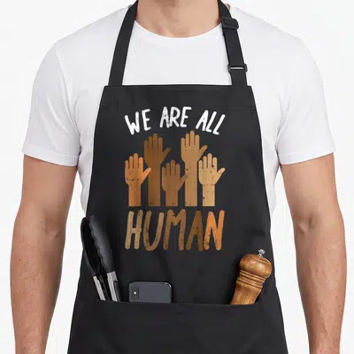 Human pride apron - Five raised hands in brown tan and white with text We Are All Human — shown on a black cotton canvas apron against a white background