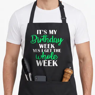 Week whole birthday apron - Stacked white and green text reading IT'S MY Birthday WEEK YES I GET THE whole WEEK — shown on a black cotton canvas apron against a white background