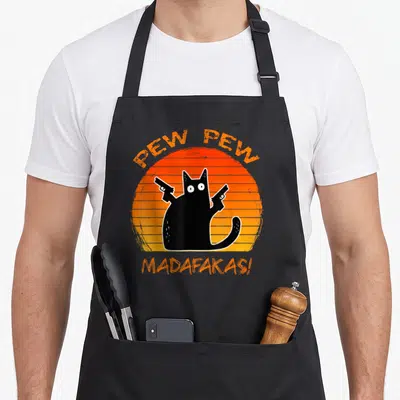 Gun cat apron - Black cat holding two guns in front of orange striped sun with arched text above and below — shown on a black cotton canvas apron against a white background