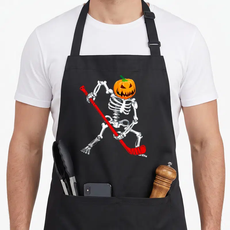 Hockey halloween apron - Cartoon skeleton with pumpkin head holding red hockey stick — shown on a black cotton canvas apron against a white background