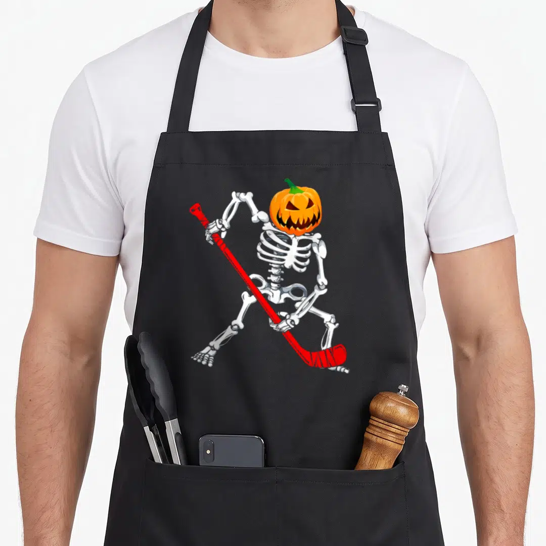 Hockey halloween apron - Cartoon skeleton with pumpkin head holding red hockey stick — shown on a black cotton canvas apron against a white background