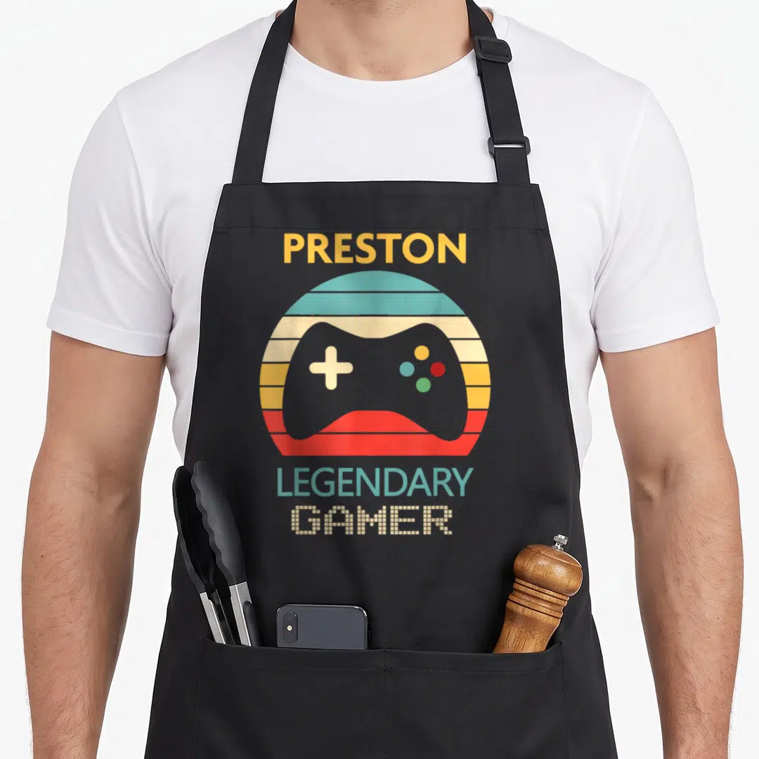 Preston gamer apron - Retro game controller in striped circle with Preston Legendary Gamer text — shown on a black cotton canvas apron against a white background