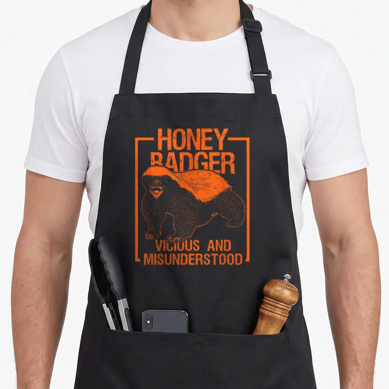 Cool honey badger apron - Orange distressed honey badger illustration with text Vicious and Misunderstood inside — shown on a black cotton canvas apron against a white background