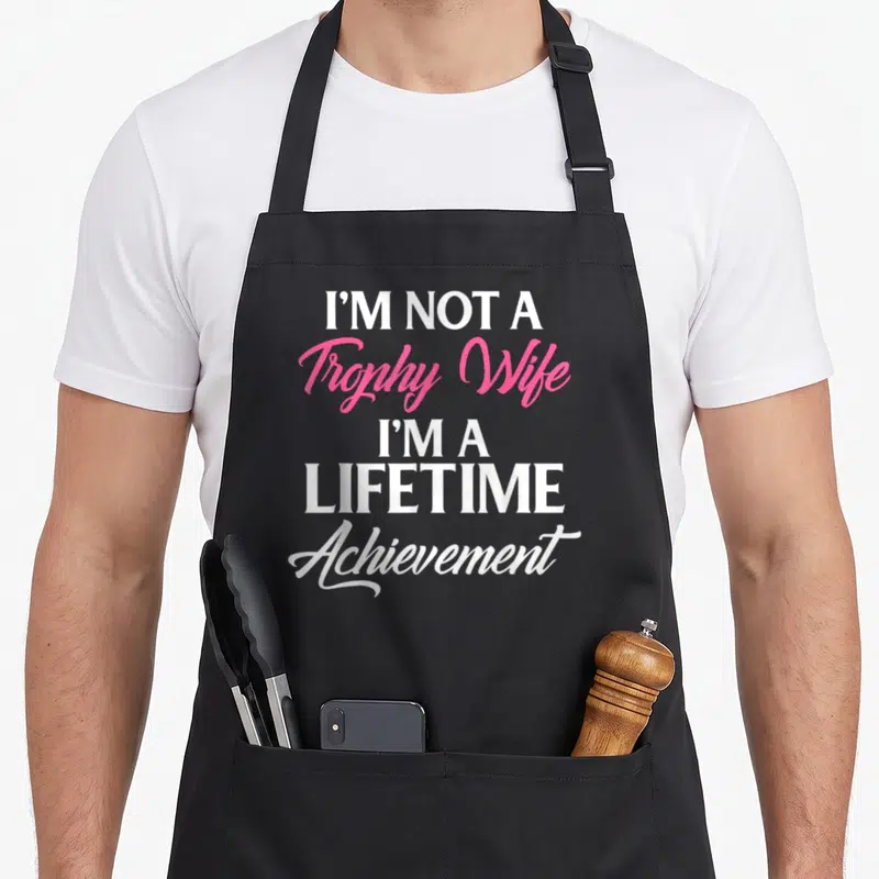 Gaming trophy-wife aprons - Stacked white and pink text reading I'm not a Trophy Wife I'm a lifetime achievement — shown on a black cotton canvas apron against a white background