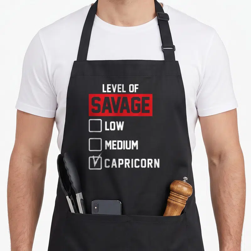 Level savage apron - Checklist graphic with Level of Savage Low Medium Capricorn text and checked box — shown on a black cotton canvas apron against a white background