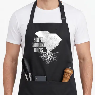 South patriotic apron - White distressed South Carolina map with roots and stacked text reading SOUTH CAROLINA ROOTS — shown on a black cotton canvas apron against a white background