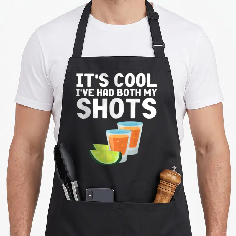 Both shot apron - Stacked white text above two orange shot glasses and two green lime wedges — shown on a black cotton canvas apron against a white background