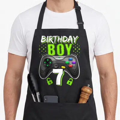 Birthday controller video game apron - Game controller with number 7, two open locks, and stacked BIRTHDAY BOY text — shown on a black cotton canvas apron against a white background