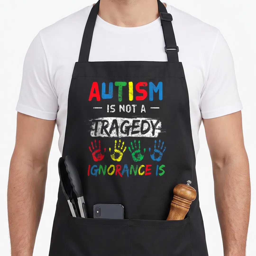 Tragedy inclusion awareness apron - Stacked text reading AUTISM IS NOT A TRAGEDY IGNORANCE IS with four colorful — shown on a black cotton canvas apron against a white background