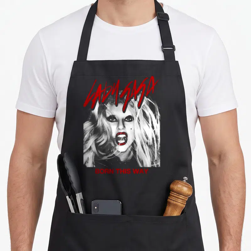 Cover novelty apron - Distressed woman portrait with red LADY GAGA and BORN THIS WAY text on grey background — shown on a black cotton canvas apron against a white background