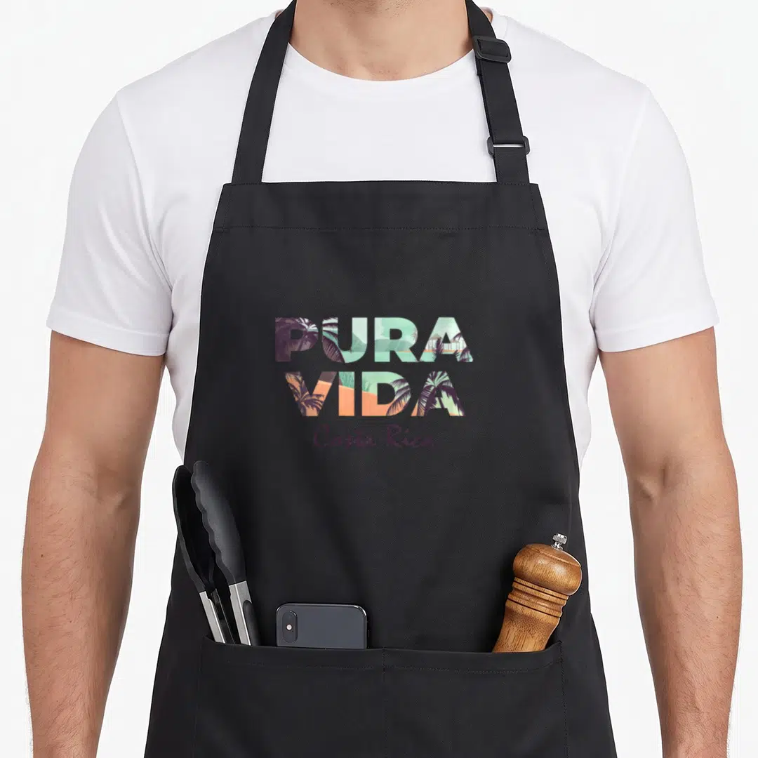 Pura colorful aprons - Stacked PURA VIDA letters containing tropical landscape in purple orange blue and green — shown on a black cotton canvas apron against a white background