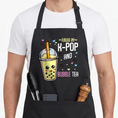 Pop travel aprons - Cute cartoon bubble tea cup with face next to stacked text FUELED BY K-POP AND BUBBLE TEA — shown on a black cotton canvas apron against a white background