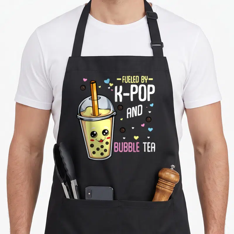 Pop travel aprons - Cute cartoon bubble tea cup with face next to stacked text FUELED BY K-POP AND BUBBLE TEA — shown on a black cotton canvas apron against a white background