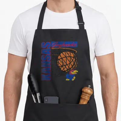 Basketball 90s apron - Vertical KANSAS text, horizontal Jayhawks script, orange basketball in net, and small bird — shown on a black cotton canvas apron against a white background