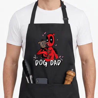 Heroe dog apron - Cartoon red character holding brown dog with DOG DAD text and hearts — shown on a black cotton canvas apron against a white background