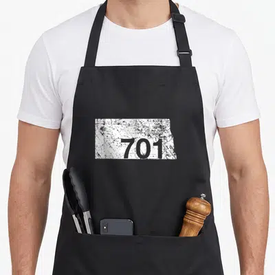 North area code apron - Distressed white North Dakota map shape with grey number 701 printed inside — shown on a black cotton canvas apron against a white background