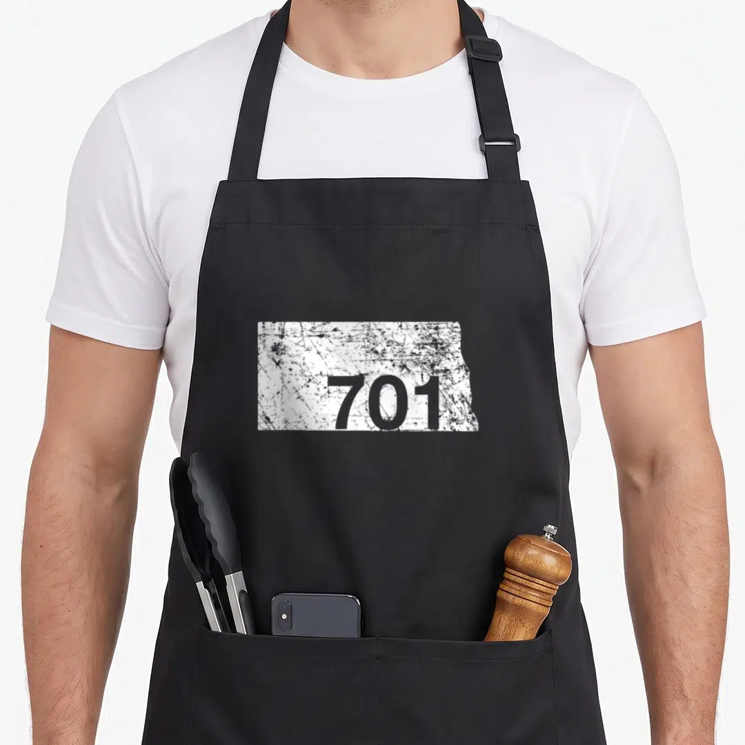 North area code apron - Distressed white North Dakota map shape with grey number 701 printed inside — shown on a black cotton canvas apron against a white background