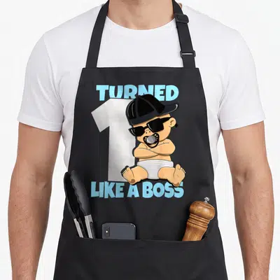 Baby bbq boss apron - Cartoon baby in sunglasses and cap sitting before number one with blue text — shown on a black cotton canvas apron against a white background