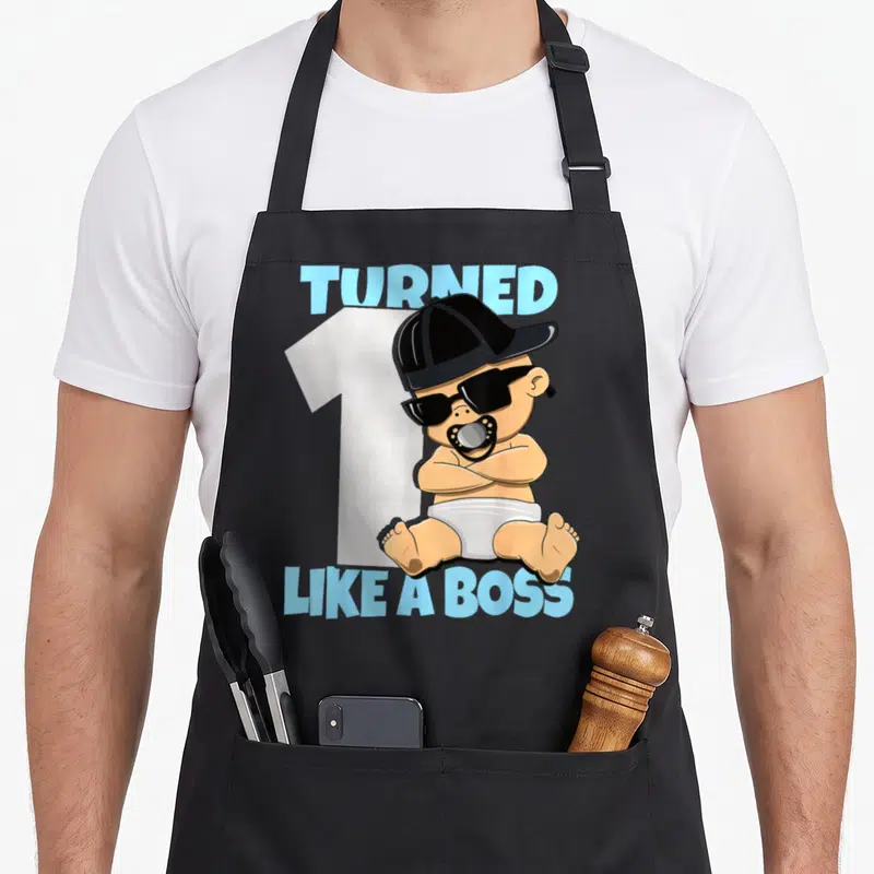 Baby bbq boss apron - Cartoon baby in sunglasses and cap sitting before number one with blue text — shown on a black cotton canvas apron against a white background