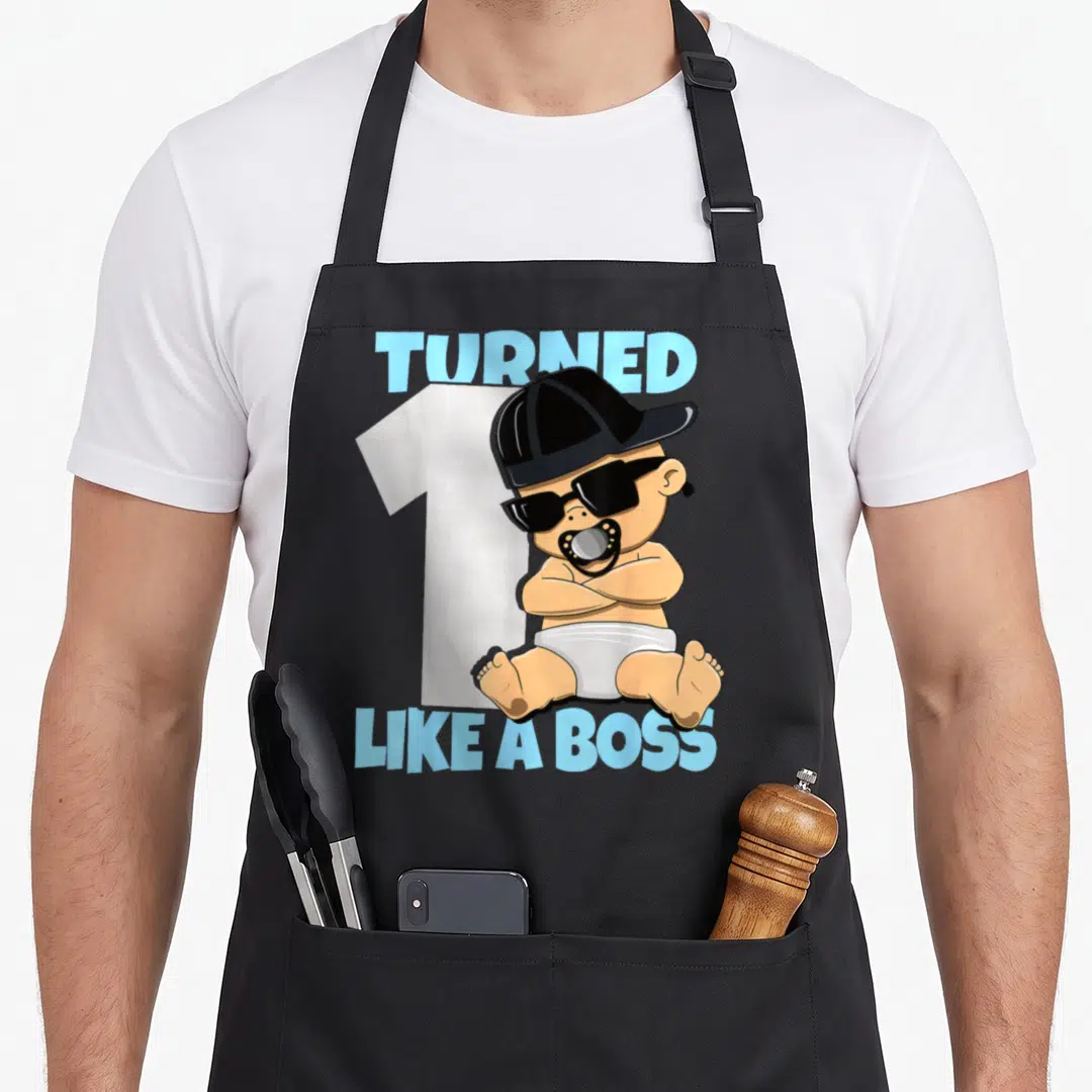 Baby bbq boss apron - Cartoon baby in sunglasses and cap sitting before number one with blue text — shown on a black cotton canvas apron against a white background