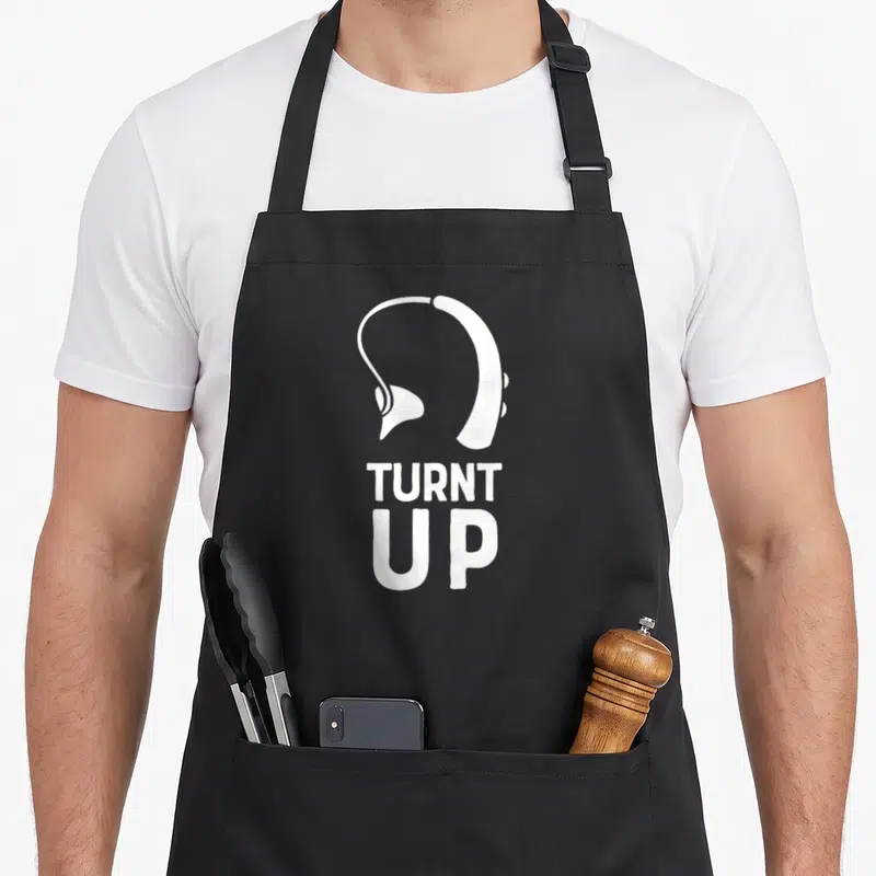 Turnt pride apron - White headphone silhouette above stacked TURNT UP text on black background — shown on a black cotton canvas apron against a white background