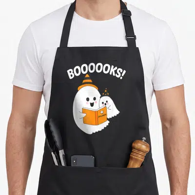 Ghostly halloween apron - Two cartoon ghosts in hats holding an orange book under arched BOOOOOKS text — shown on a black cotton canvas apron against a white background