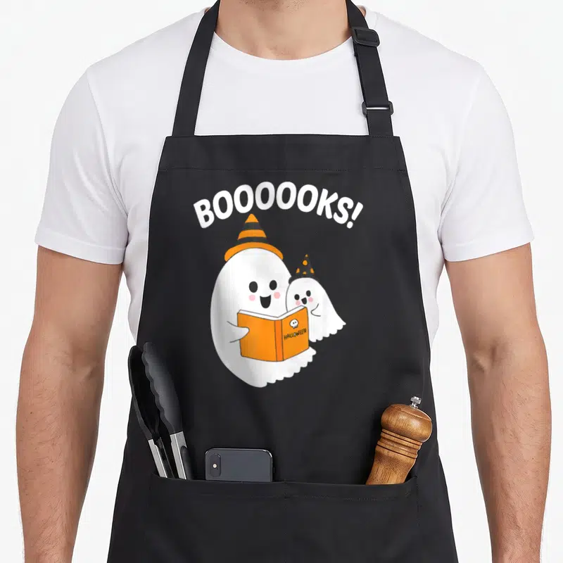 Ghostly halloween apron - Two cartoon ghosts in hats holding an orange book under arched BOOOOOKS text — shown on a black cotton canvas apron against a white background