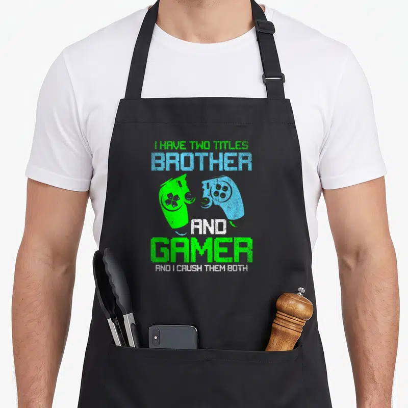 Gamer brother video game apron - Stacked text I HAVE TWO TITLES BROTHER GAMER with two facing green and blue — shown on a black cotton canvas apron against a white background