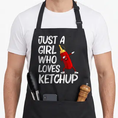 Merchandise tomato apron - Stacked white text next to a standing red cartoon ketchup bottle with white gloves and shoes — shown on a black cotton canvas apron against a white background