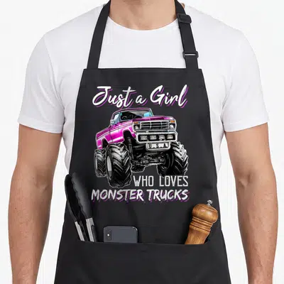 Funny monster birthday truck apron - Pink monster truck illustration with Just a Girl Who Loves Monster Trucks text — shown on a black cotton canvas apron against a white background