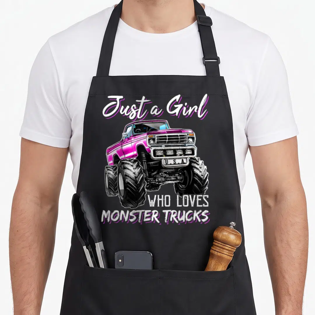 Funny monster birthday truck apron - Pink monster truck illustration with Just a Girl Who Loves Monster Trucks text — shown on a black cotton canvas apron against a white background