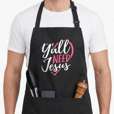 Jesus religious christian apron - Y'all NEED Jesus text in white and pink inside a heart outline — shown on a black cotton canvas apron against a white background