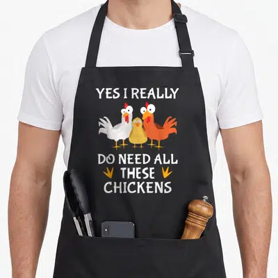 Farmer do i need an apron - Three cartoon chickens in white orange and yellow with text and small feet icons — shown on a black cotton canvas apron against a white background