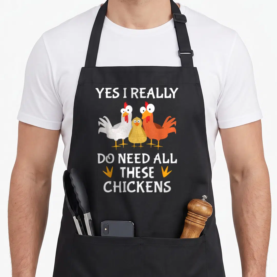 Farmer do i need an apron - Three cartoon chickens in white orange and yellow with text and small feet icons — shown on a black cotton canvas apron against a white background