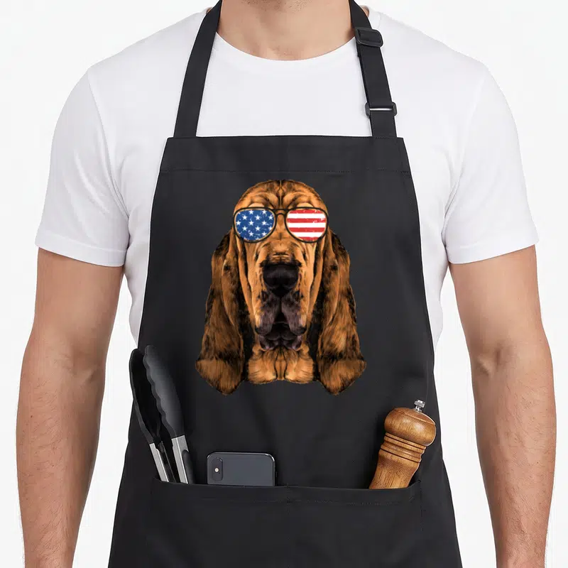 Bloodhound american flag apron - Realistic bloodhound head wearing sunglasses with red white and blue flag pattern — shown on a black cotton canvas apron against a white background