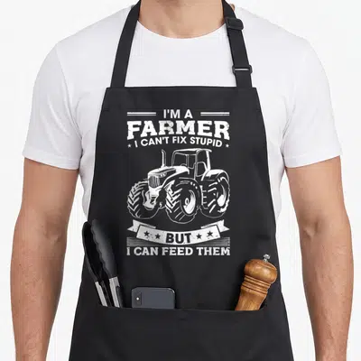 Tractor farming apron - Stacked text reading I'm a farmer I can't fix stupid but I can feed them with a tractor — shown on a black cotton canvas apron against a white background